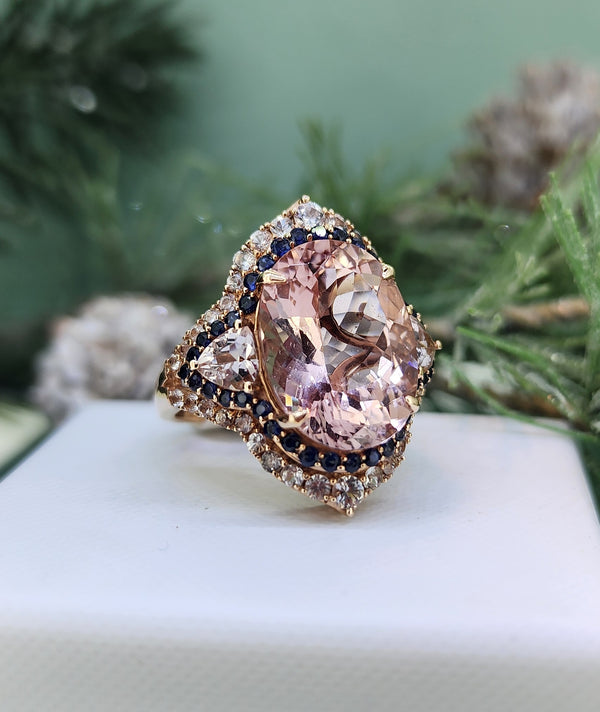 Crafted in warm 14k rose gold, this exquisite ring showcases a luminous 15x10mm morganite center stone, softly blushing with peach and rose tones. The morganite is framed by a delicate halo of natural blue sapphires, adding depth and contrast, while brilliant white sapphires complete the design with a graceful sparkle. The result is a richly detailed ring that feels both romantic and timeless, with a subtle nod to vintage elegance