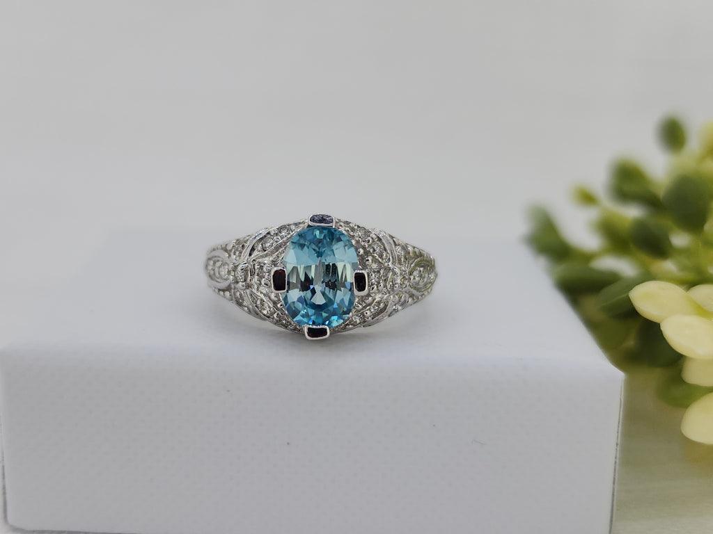 Beautifully detailed and thoughtfully crafted, this ring features an 8.00 × 5.70mm oval faceted blue zircon, prized for its vibrant blue color and exceptional brilliance. The center stone is accented by round brilliant-cut natural mined diamonds totaling .15 carat total weight, adding refined sparkle throughout the design.

Crafted in 14K white gold, this vintage-inspired ring showcases intricate detailing and excellent workmanship. Measuring 10mm across the top and tapering to a 2.34mm shank, it offers a b