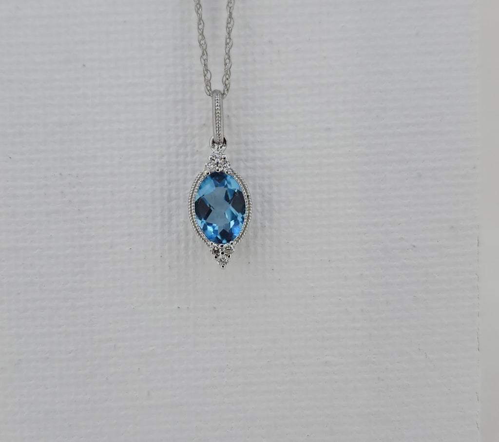 Elegant and softly detailed, this pendant features a 7×5mm oval Swiss blue topaz with luminous color and graceful faceting. The center stone is accented by .06 carat total weight of natural mined diamonds, adding delicate sparkle and balance to the design.

Crafted in 14K white gold, the pendant is finished with milgrain detailing, a refined beaded edge that adds texture and depth. Measuring 17.00mm in length and 6.11mm in width, it is suspended from an 18-inch white gold Singapore chain and secured with a 