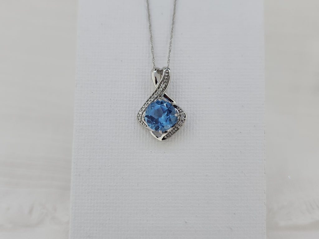 Elegant with a bit of modern flare, this pendant features an 8mm round faceted Swiss blue topaz, admired for its vibrant blue color and brilliant sparkle. The center stone is accented by .10 carat total weight of natural mined round brilliant diamonds, adding refined shimmer and contrast. Crafted in 14K white gold, the pendant measures 19.00mm in length and 13.28mm in width and is hung from an 18-inch white gold Singapore chain with a spring ring clasp. With a total weight of 2.0 grams including the chain 