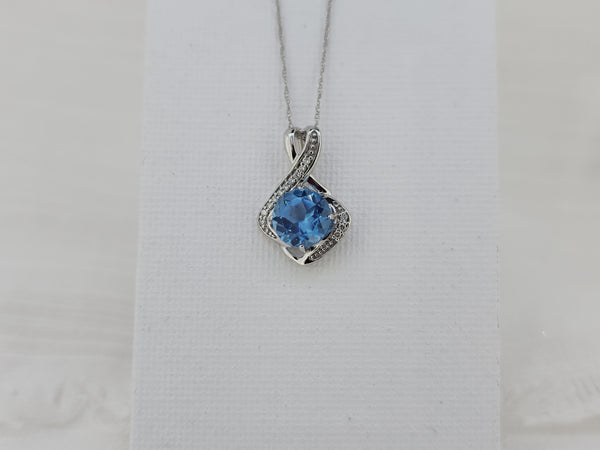 Elegant with a bit of modern flare, this pendant features an 8mm round faceted Swiss blue topaz, admired for its vibrant blue color and brilliant sparkle. The center stone is accented by .10 carat total weight of natural mined round brilliant diamonds, adding refined shimmer and contrast. Crafted in 14K white gold, the pendant measures 19.00mm in length and 13.28mm in width and is hung from an 18-inch white gold Singapore chain with a spring ring clasp. With a total weight of 2.0 grams including the chain 