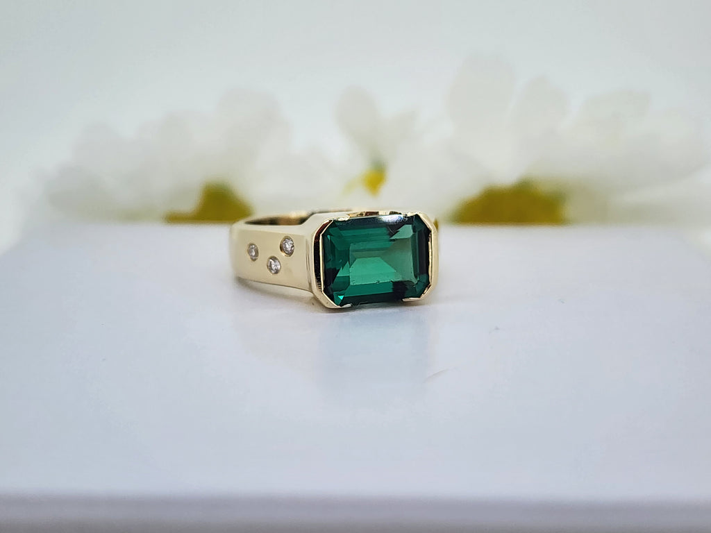 The vibrant color of this created Emerald is the color you dream that perfect Emerald to be! This is a beautifully crafted piece weighing in at 8.50 grams. The center stone measures 10x8mm and has a few small chips and light abrasion. On each side of the center stone are (3) three round brilliant cut natural mined diamonds that have a combined total weight of .11cttw. G/H in color. SI goods. The ring measures 8.32mm at the top and tapers to a 4.60mm  shank. 