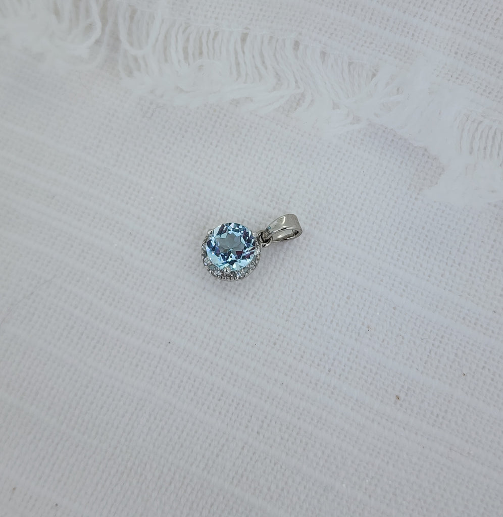 Delicate yet brilliantly detailed, this pendant features a natural 6mm round diamond-cut aquamarine, The center stone is beautifully framed by a halo of nine round brilliant-cut natural mined diamonds, with a combined .09 carat total weight. Crafted in 14K white gold, the refined setting enhances the aquamarine’s cool tone while maintaining a timeless, versatile look. The pendant measures 13.28mm in length including the bail and 7.47mm in width. Sold without a chain