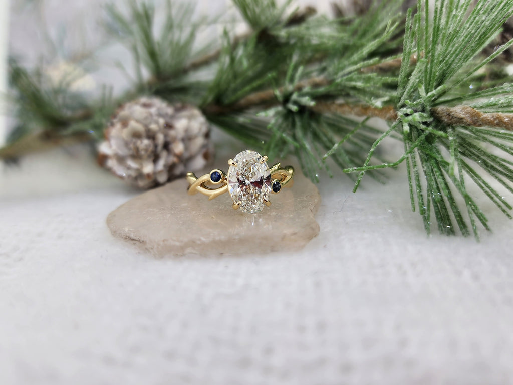 14k yellow gold branch mounting set with a beautiful 2.13ct oval Lab grown diamond. The diamond is F in color and VS1 in clarity. The diamond is set in a four prong head with a 2.00mm natural blue sapphire bezel set on each side of the center diamond. USA finger size 7